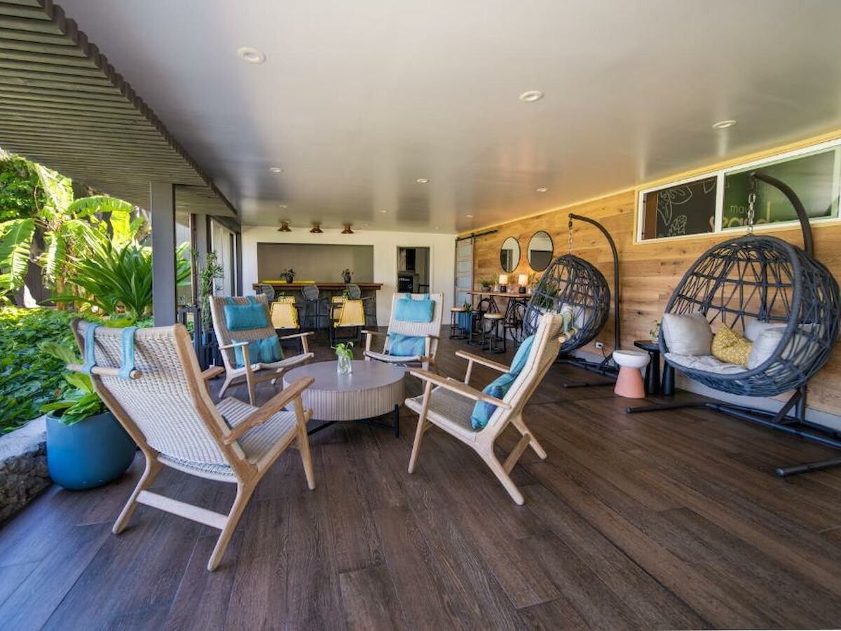 A modern patio area with wooden flooring, various chairs, hanging egg chairs, a coffee table, and lush greenery in the background.