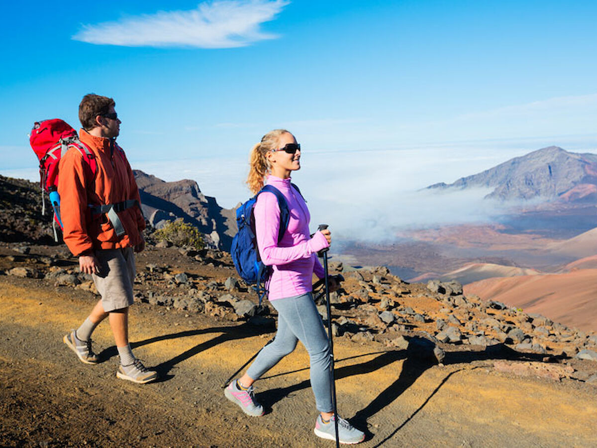 Two hikers, one man and one woman, walk along a rocky trail with mountainous scenery in the background under a clear blue sky.
