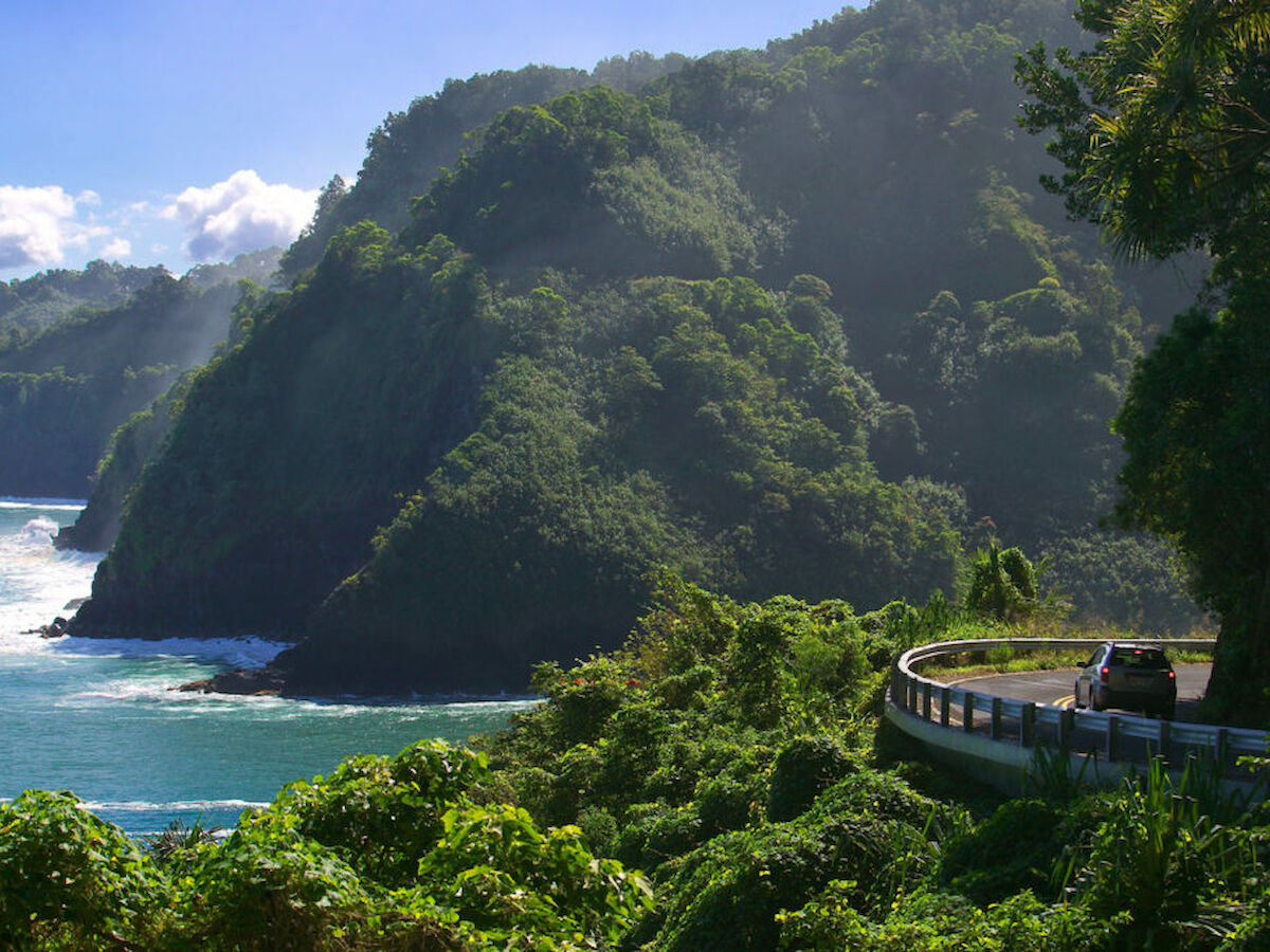 A coastal road winds through lush green hills with a car driving along it, adjacent to the ocean with waves hitting the shore, completing the scene.