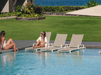 Two people relax by a pool with lounge chairs, lush greenery, and ocean in the background under a clear sky.