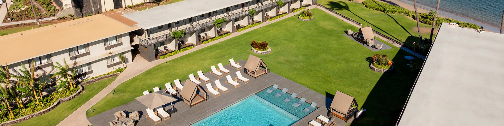 Aerial view of a coastal resort with a pool, lounge chairs, buildings, and palm trees, set against a mountainous backdrop.