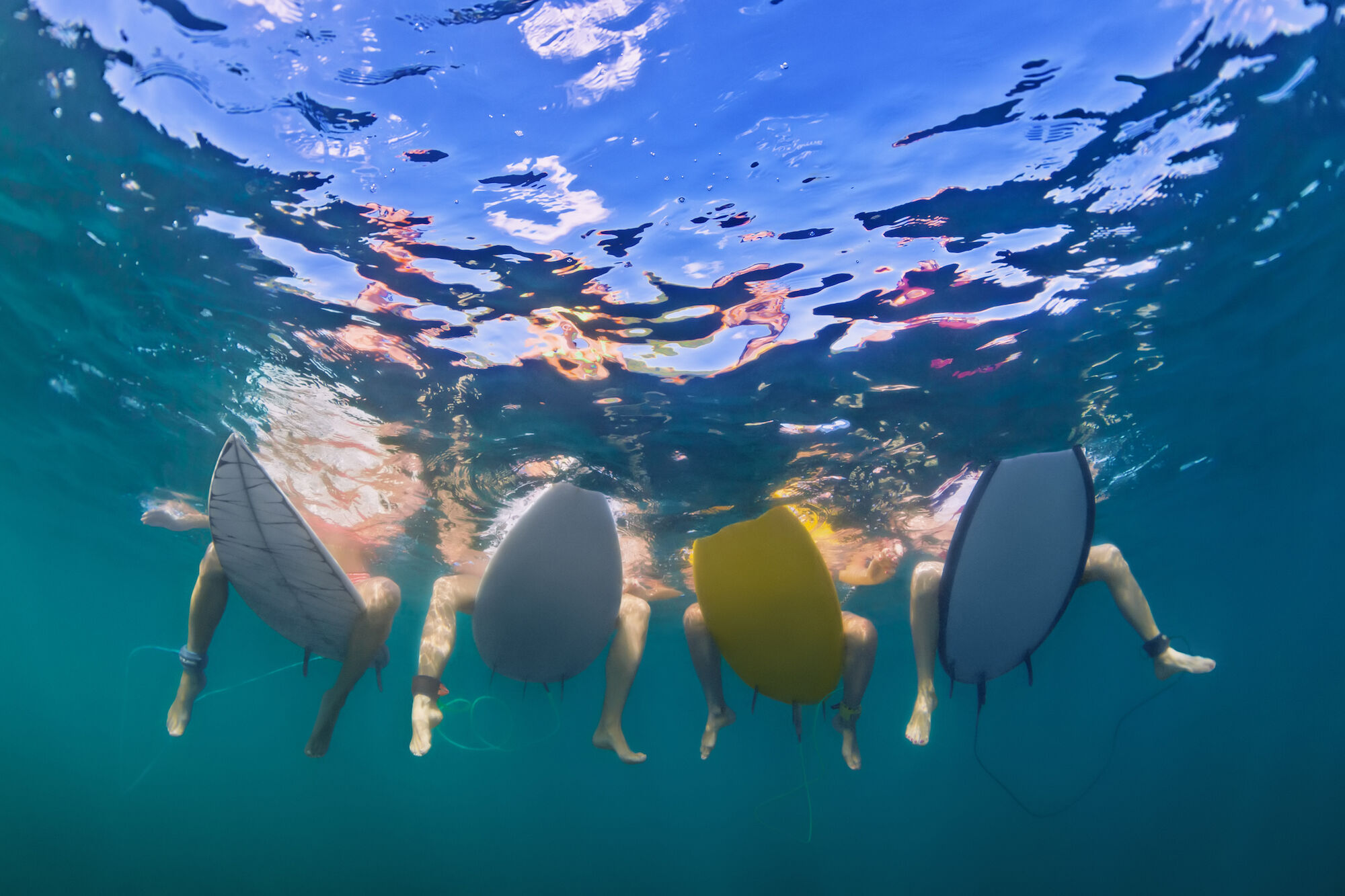 This image shows four people with surfboards sitting in the water, viewed from below the surface of the water.