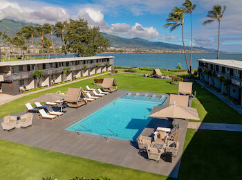 A resort with a central pool, surrounded by lounge chairs, buildings, palm trees, and a scenic view of mountains and the sea.