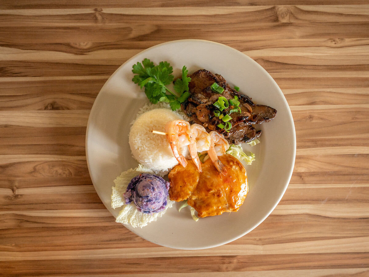 The image shows a plate with rice, shrimp, grilled meat, sauce-covered item, garnished with cilantro, on a wooden table.