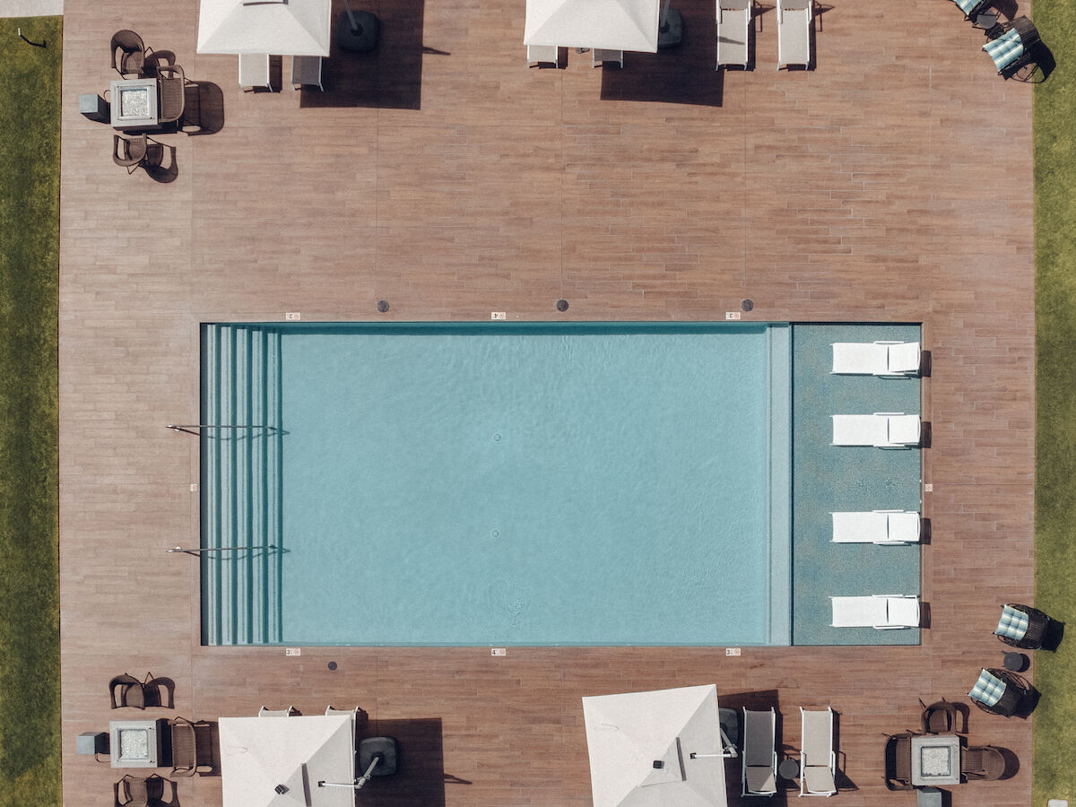 An overhead view of a rectangular swimming pool surrounded by patio chairs, umbrellas, and green grass. Four cabanas with loungers are visible.