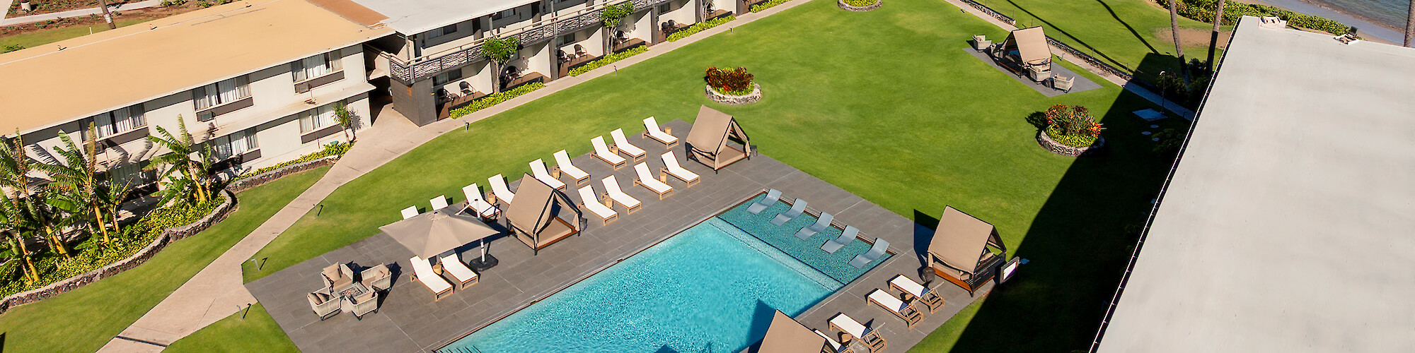 Aerial view of a hotel by the beach with a pool, lounge chairs, and a mountain backdrop under a partly cloudy sky.