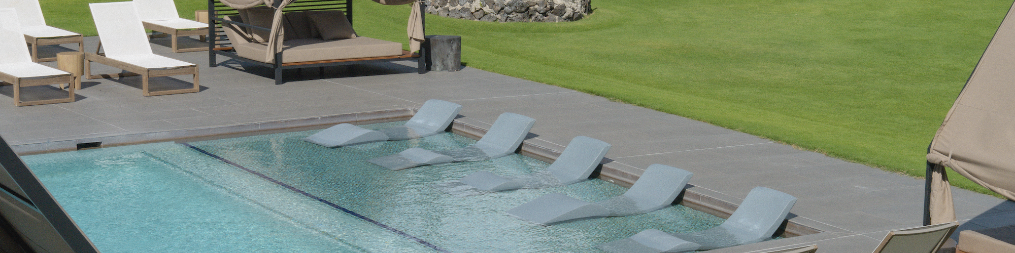A pool with loungers and sunshades on a patio, surrounded by green lawns and a row of rooms in the background.
