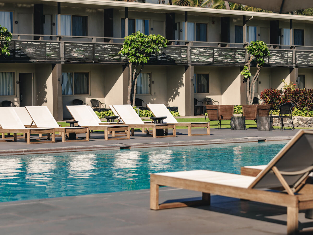 The image shows a swimming pool with wooden loungers in front of a resort building lined with balconies and greenery.