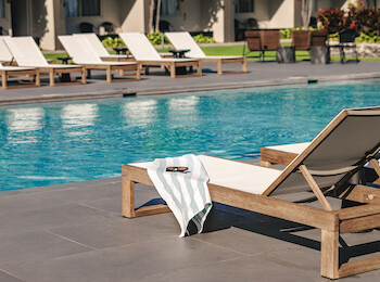 A swimming pool scene with wooden lounge chairs, one with a towel, on a tiled deck, suggesting a relaxing resort atmosphere.