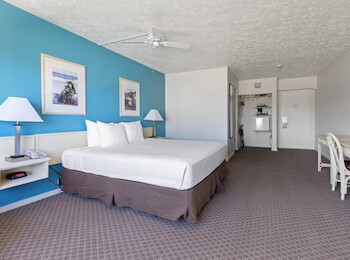 The image shows a hotel room with a bed, blue accent wall, lamps, desk, and artwork, creating a clean and minimalistic look.
