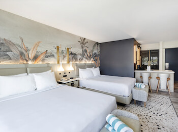 The image shows a modern hotel room with two beds, a mural wall, a sitting area, and a small bar with stools, featuring a sleek design.