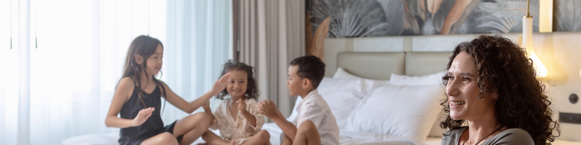 A woman sits smiling with a drink, while three children play and laugh on a bed in a bright room.