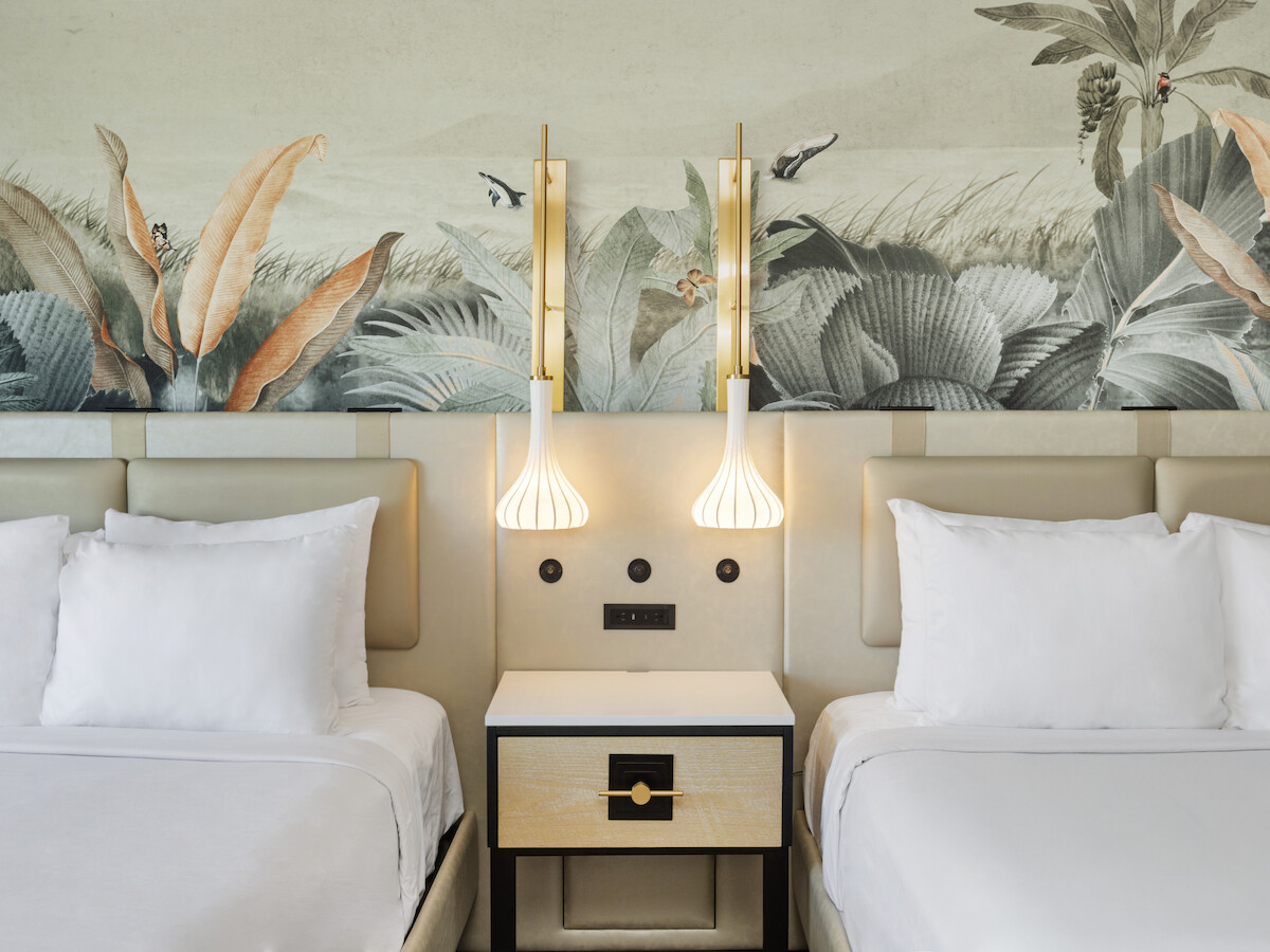 Two neatly made beds with white bedding, a nightstand, and stylish wall sconces, set against a leafy mural background.
