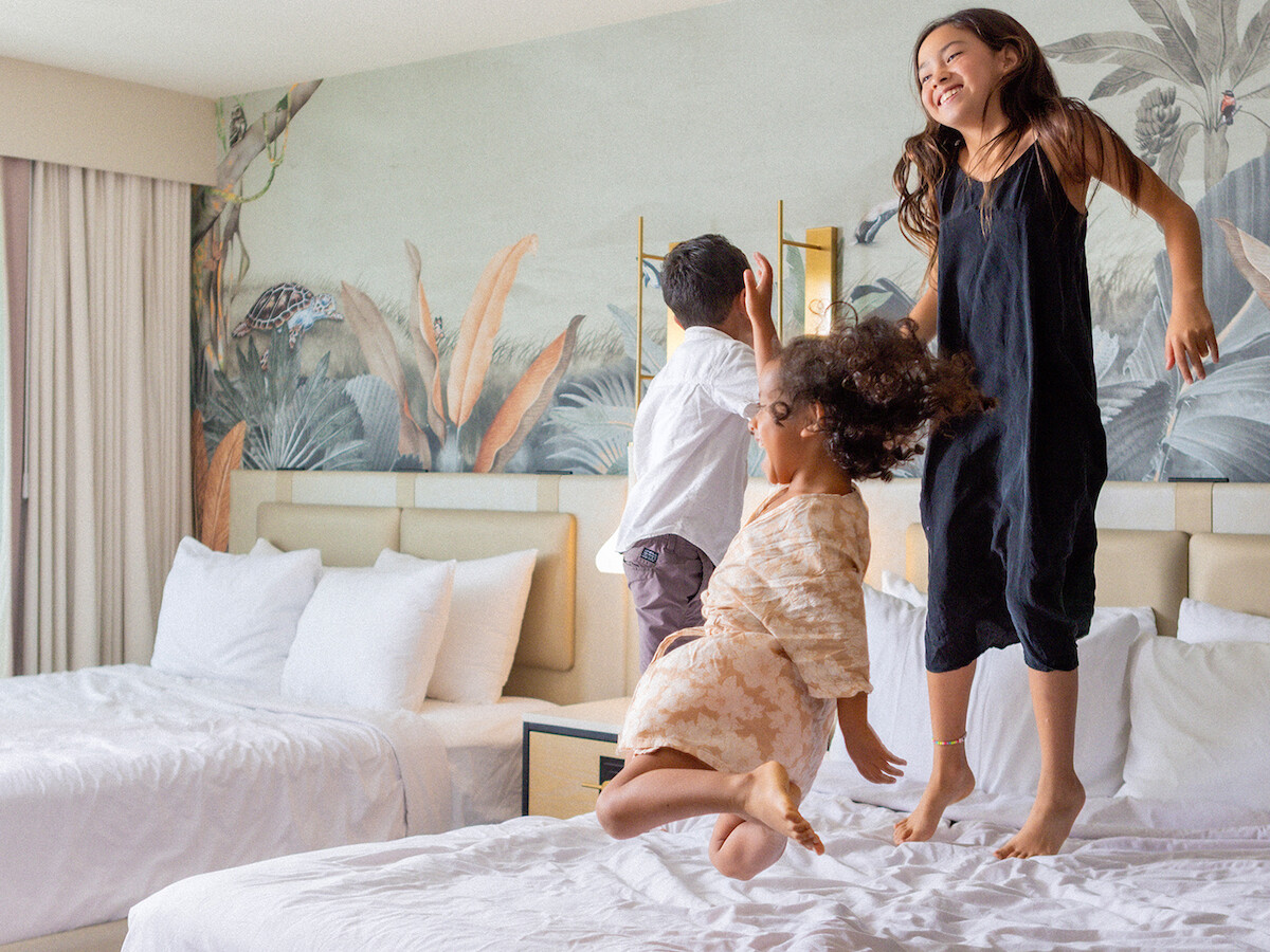 Three children are joyfully jumping on hotel beds in a room with bright decor and a nature-themed mural on the wall.