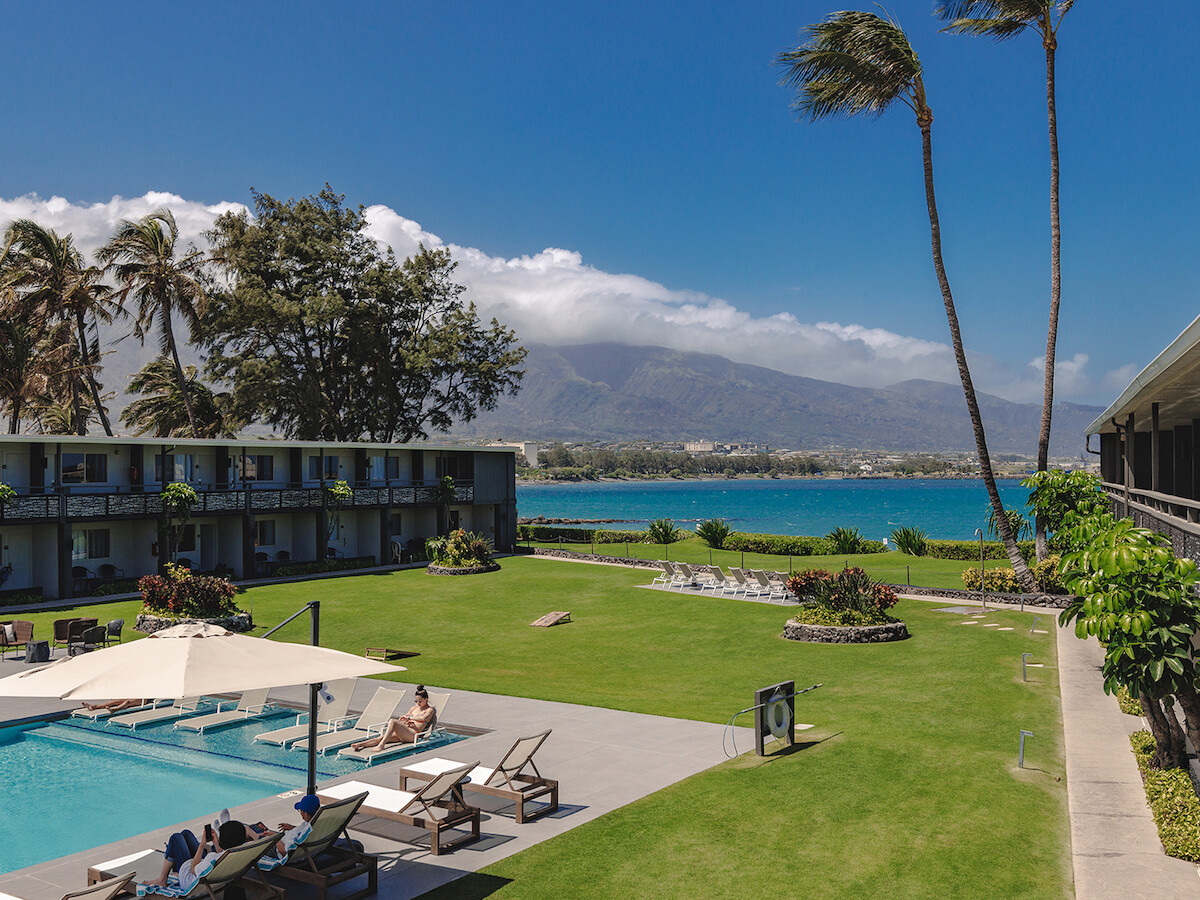 A scenic view of a resort with a pool, lounge chairs, palm trees, and a mountain and ocean backdrop under a clear blue sky.