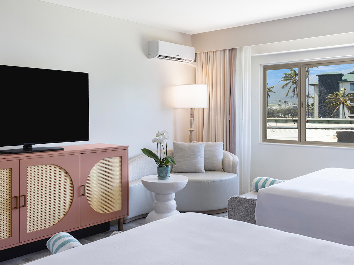 A modern hotel room with two beds, a large TV on a cabinet, a cozy seating area, and a view of palm trees through the window.