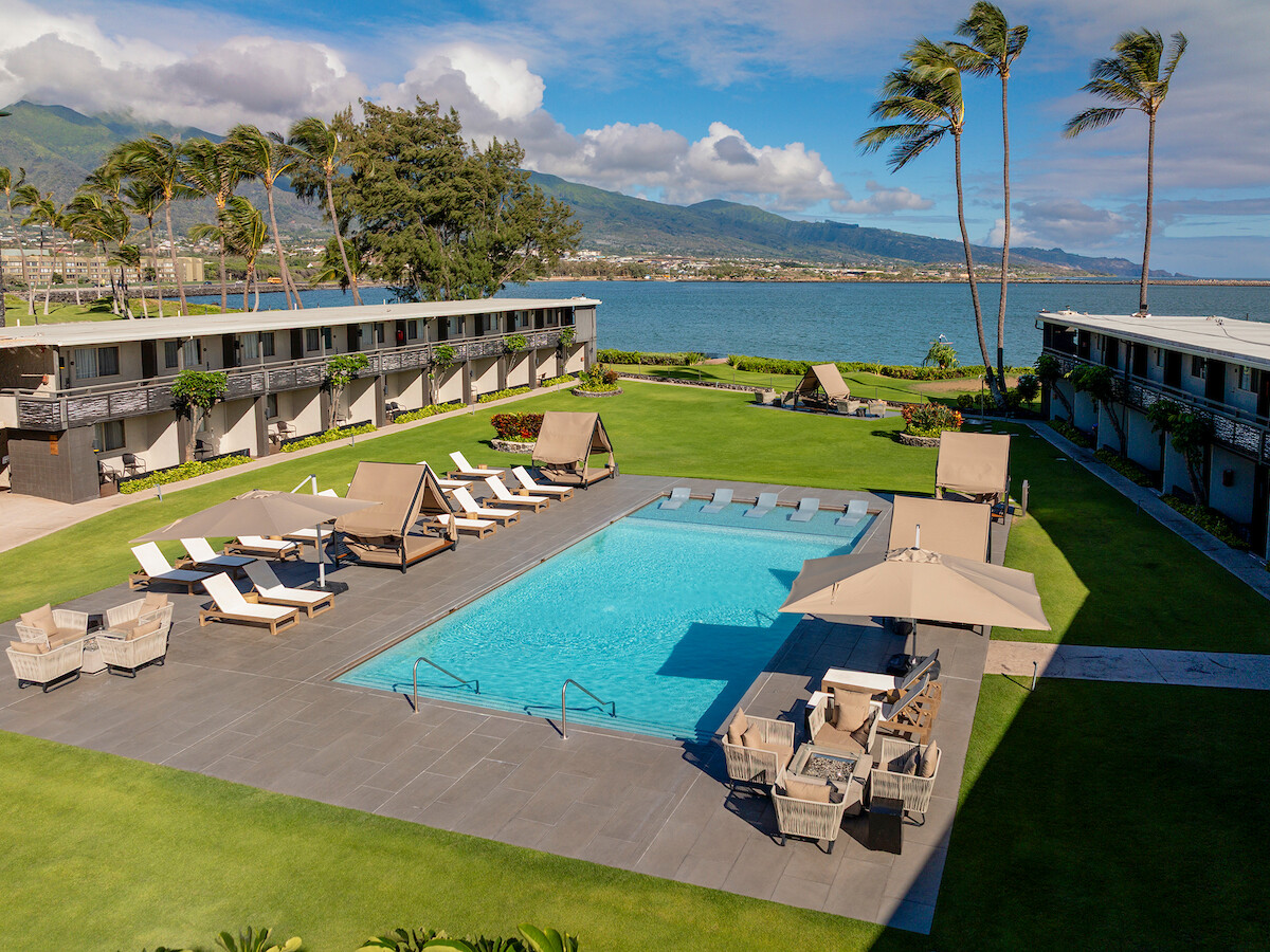 A hotel pool area with lounge chairs and umbrellas, surrounded by grassy areas and buildings, set against an ocean and mountain backdrop.