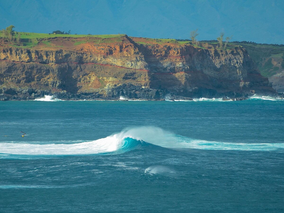 A stunning ocean scene with a wave forming in the foreground, and rugged cliffs in the background under a blue sky.