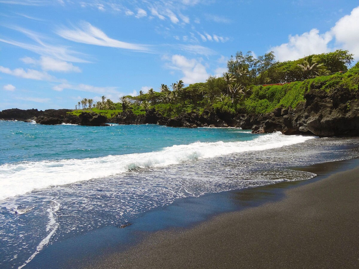A beach with black sand, clear blue water, and lush greenery under a partly cloudy sky.