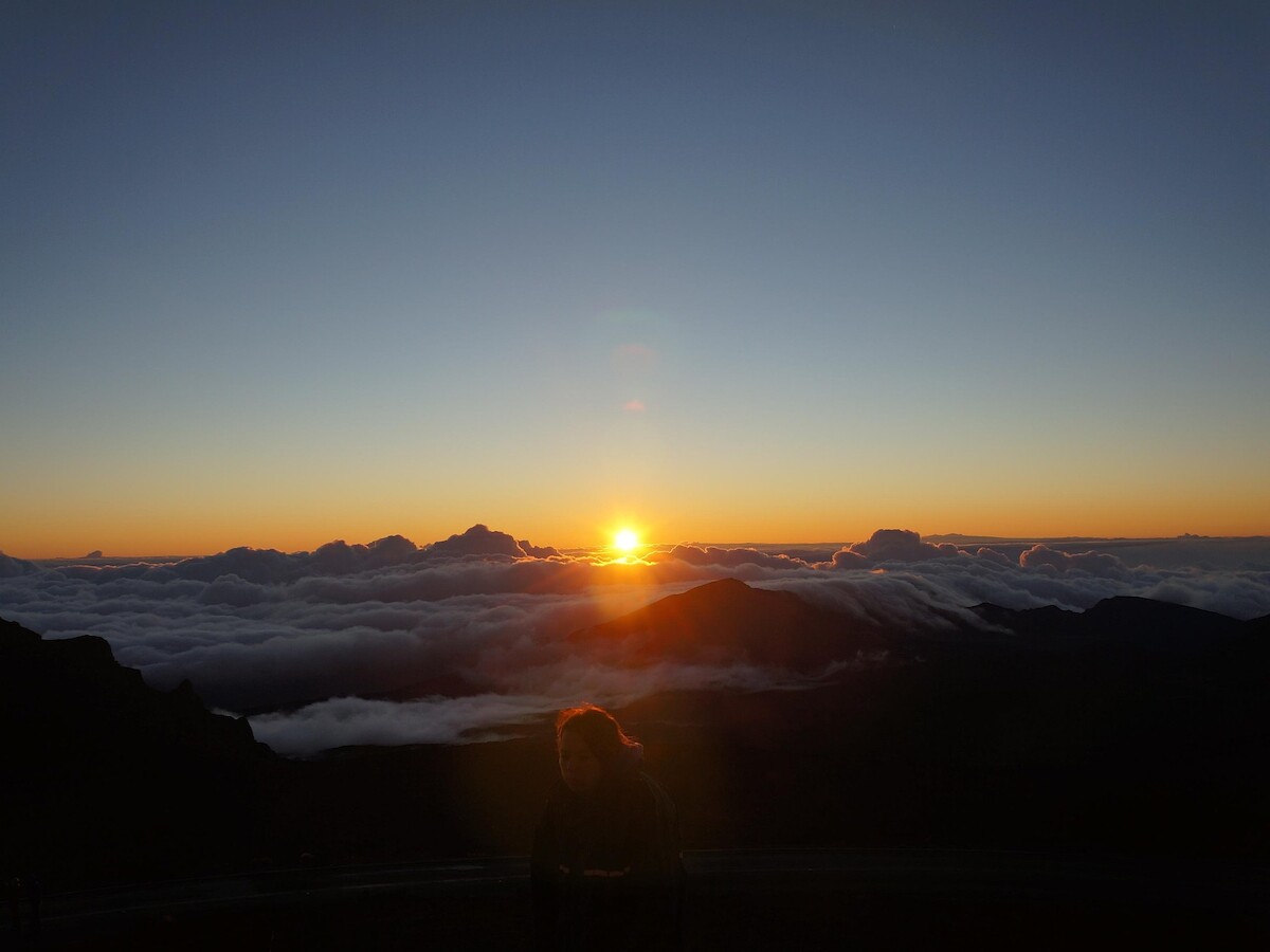 A breathtaking mountain sunrise with a silhouette of a person, clouds below, and a bright sun on the horizon.