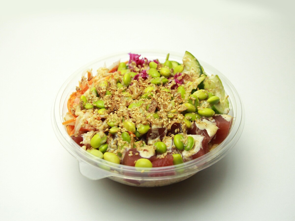 A poke bowl with edamame, cucumber, cabbage, and other fresh ingredients in a clear plastic container.
