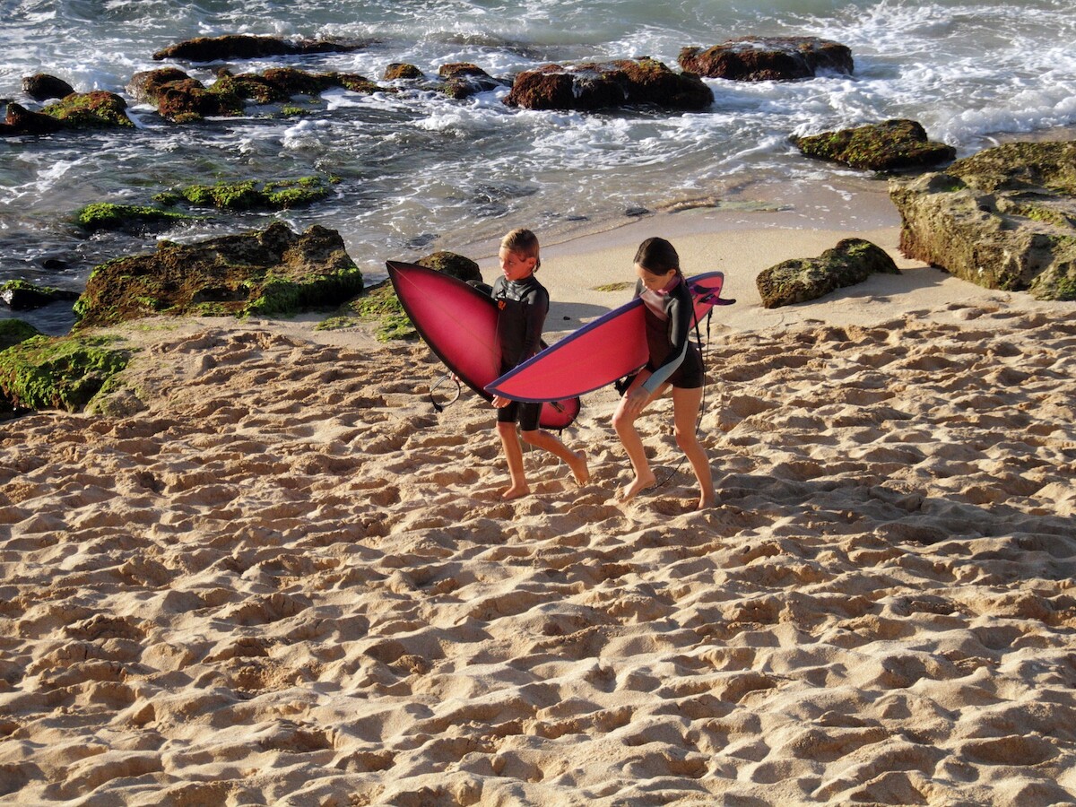 Two people are on a beach carrying red surfboards, walking towards the water with rocky areas in the background under a partly cloudy sky.