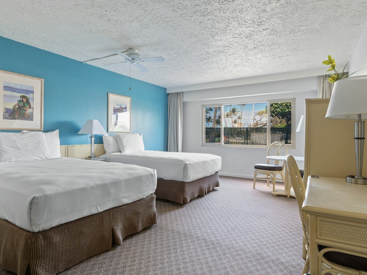 The image shows a hotel room with two beds, a ceiling fan, wall art, a window with a view, and a dresser with a lamp and mirror.