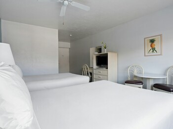 The image shows a hotel room with two beds, a TV, two chairs, a small table, and a framed picture on the wall.
