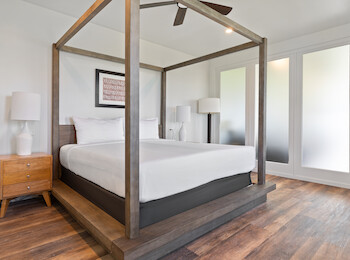 A modern bedroom with a four-poster bed, white linens, wooden nightstands, lamps, and frosted glass windows in a clean, minimal design.