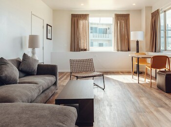 A modern living room with a gray sofa, a coffee table, a woven chair, a desk, and large windows with brown curtains.