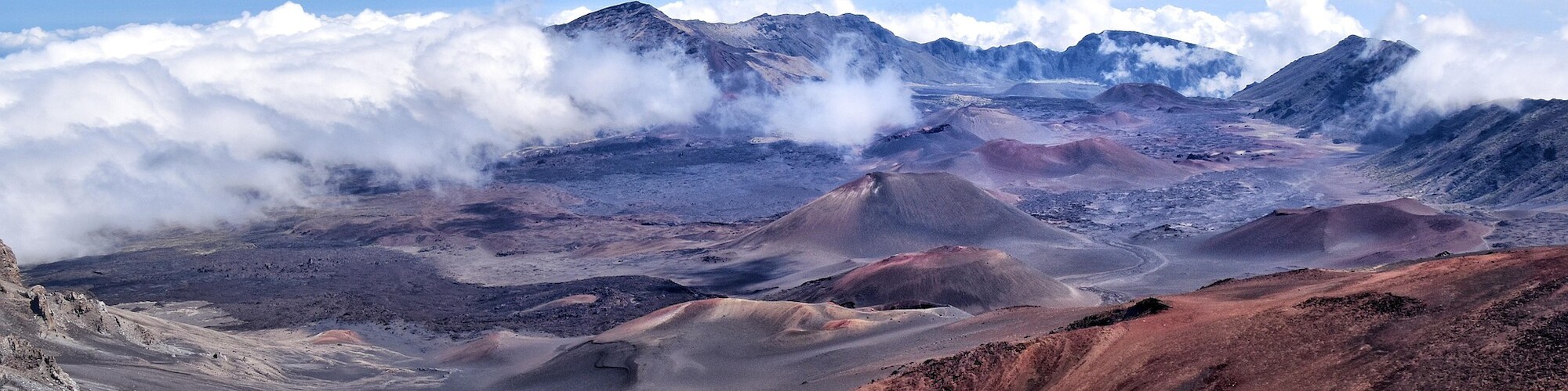 A mountainous landscape with a crater-like terrain, surrounded by clouds under a clear blue sky.
