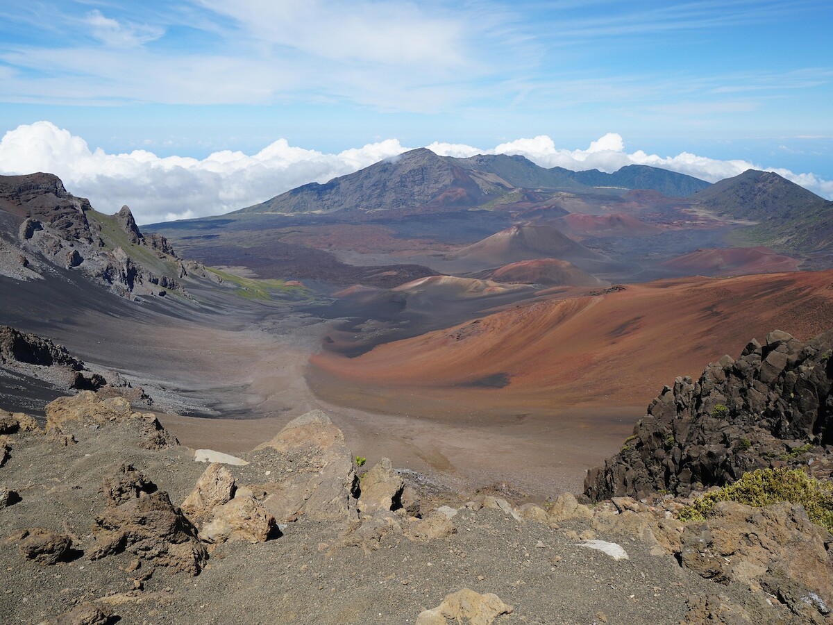 A scenic view of a volcanic landscape with colorful terrain, mountains, and a clear blue sky with some clouds in the distance.