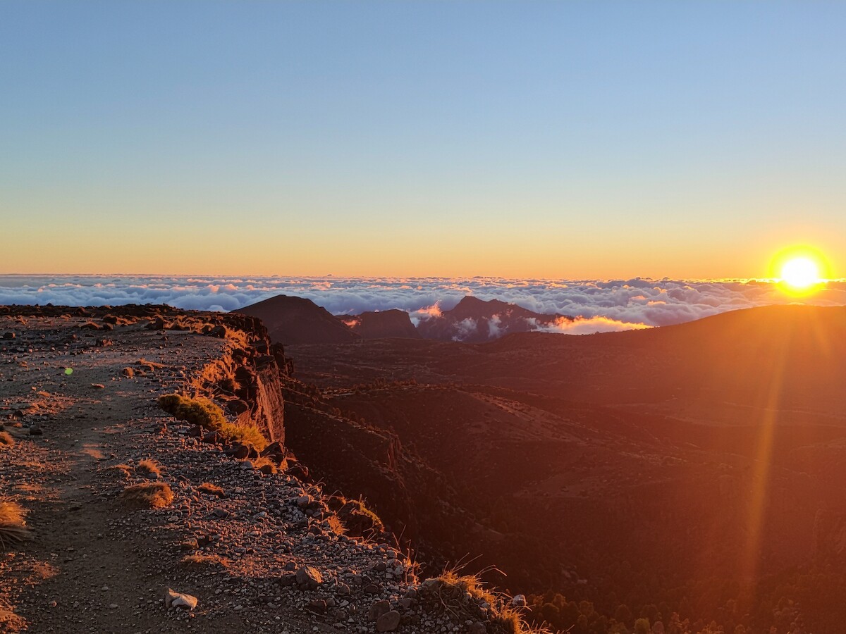 A breathtaking sunrise over a mountainous landscape, with a cliffside path and clouds visible in the distance.