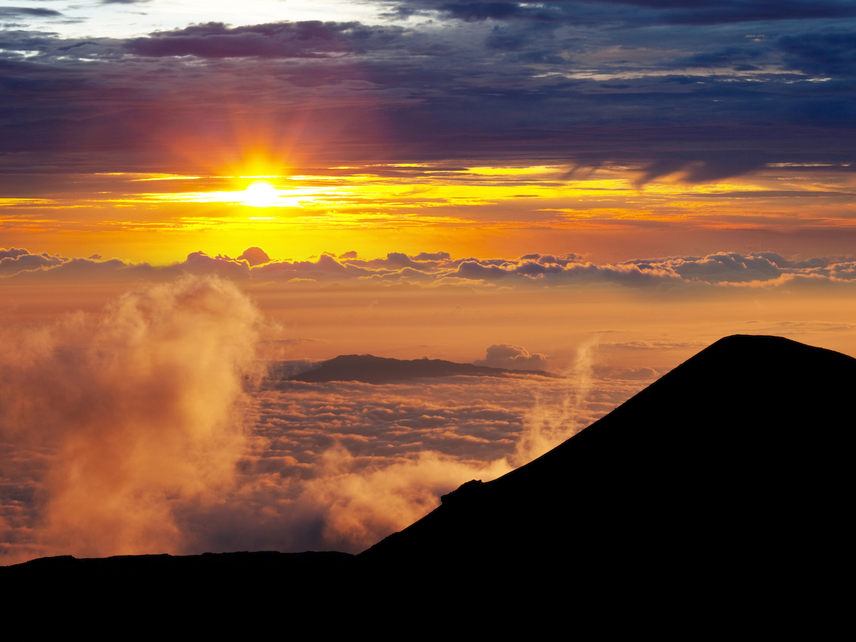A volcanic silhouette against a vibrant sunset sky, with clouds and sun rays creating a dramatic and peaceful scene.