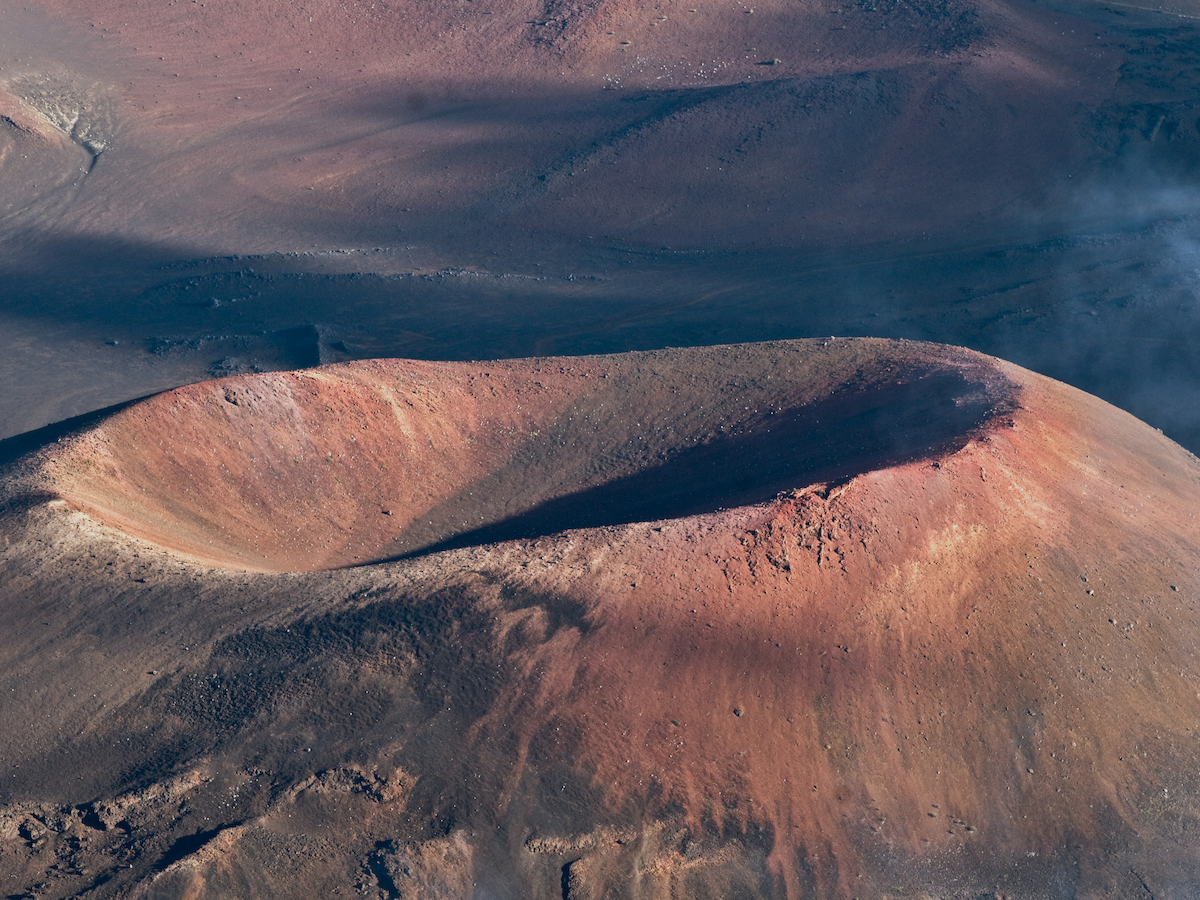 This image shows a volcanic crater with reddish-brown earth, likely part of a larger volcanic landscape under partial shadow.