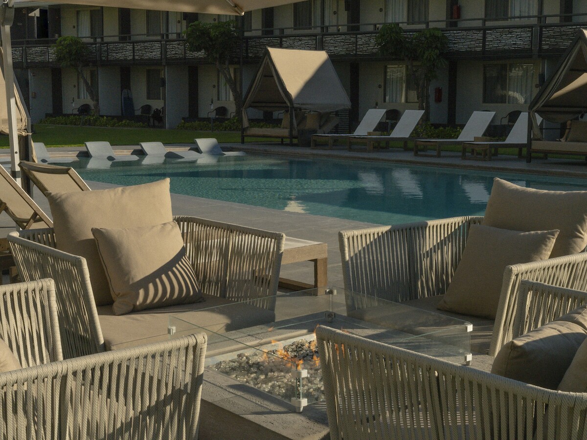 Outdoor seating with cushioned chairs around a glass table near a pool, with umbrellas and loungers in the background at a resort.