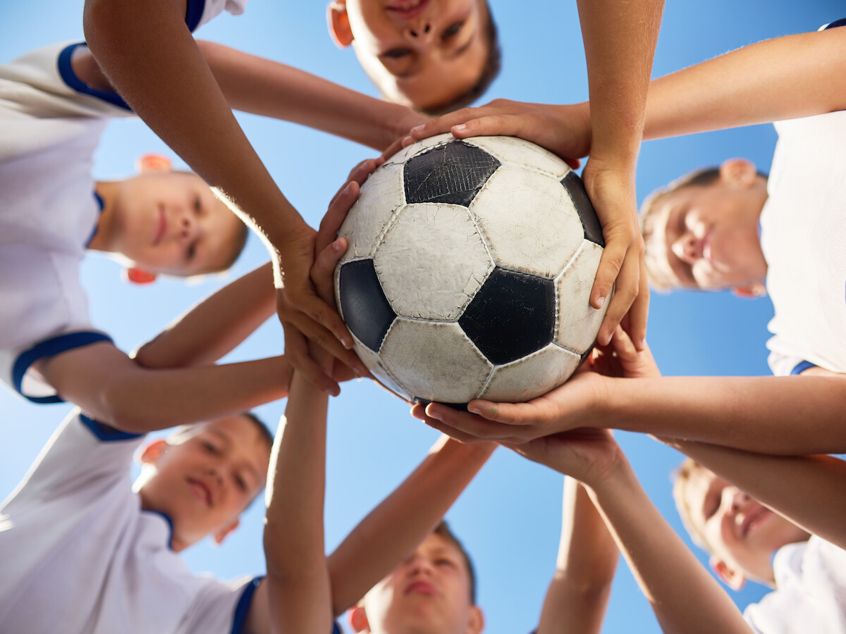 A group of kids in sports uniforms are huddled together, each with hands on a soccer ball, looking upwards with a blue sky above.
