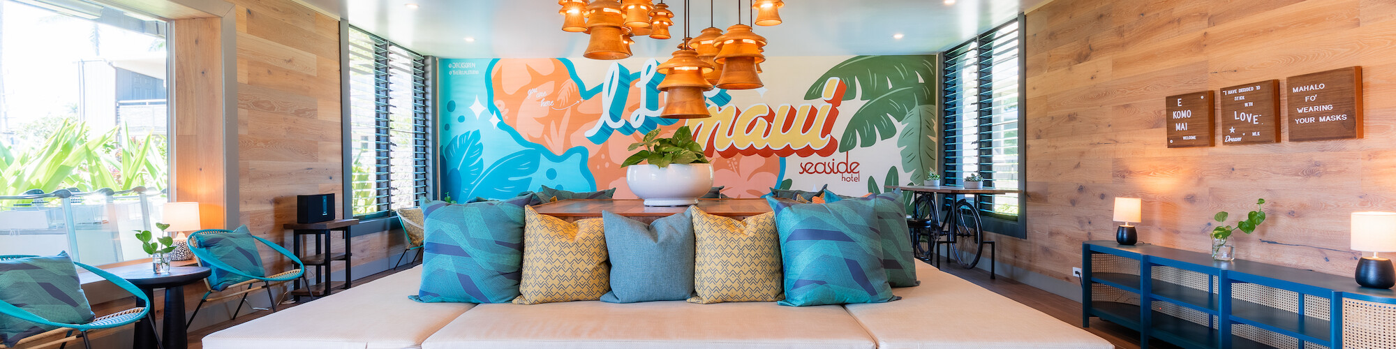 This image shows a modern lounge area with a large seating space, colorful mural, hanging lights, and cushions in a well-lit room.