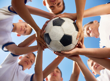 A group of kids in white shirts are holding a soccer ball together, forming a circle, with the sky visible in the background.