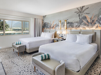 A modern hotel room with two beds, a nature-themed wall mural, stylish decor, and a window view of trees and mountains.