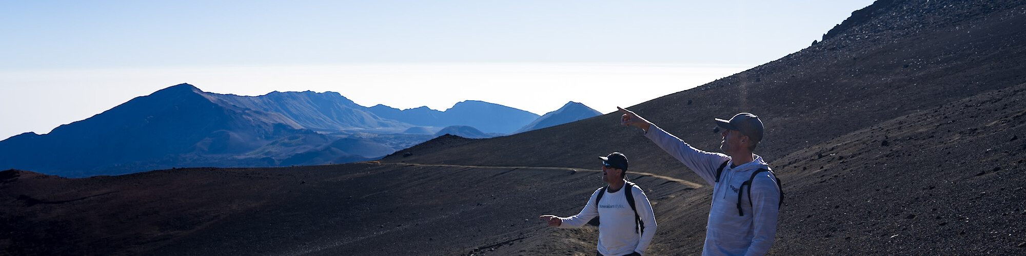 Two hikers on a barren mountain trail, one points towards the horizon, with distant peaks under bright sky.