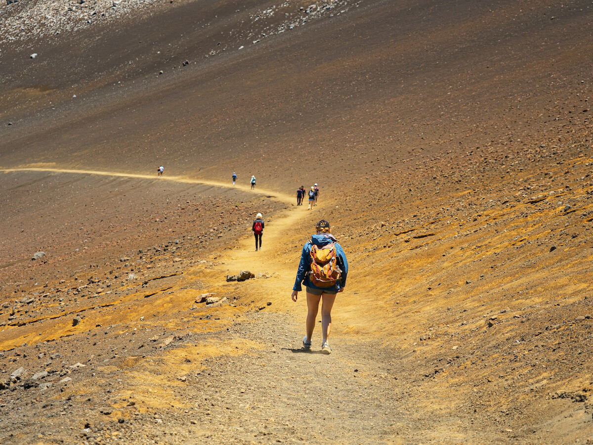 A group of hikers walking on a dusty, winding trail in a barren, rocky landscape under a clear sky, with one hiker leading the way.