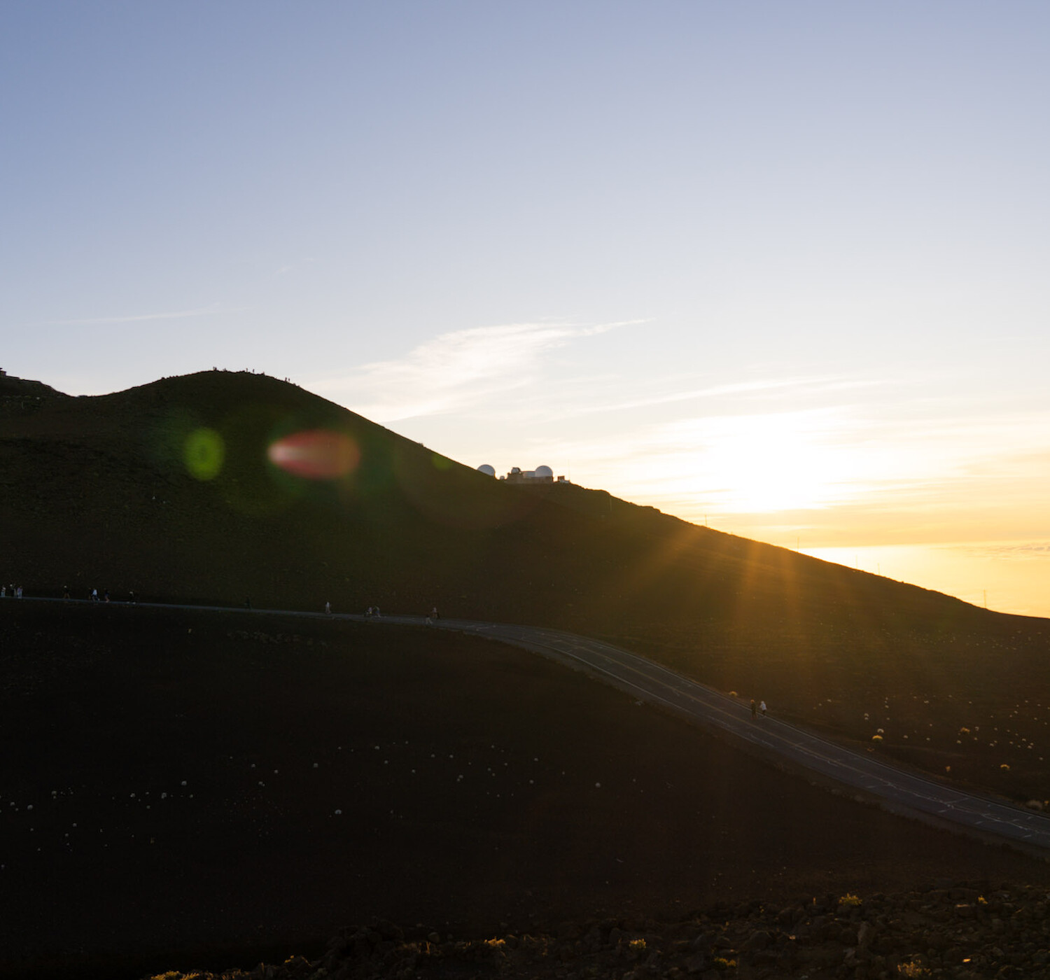 A serene sunset over a hillside with a winding road leading up, with clear skies and a soft sun glow in the background.