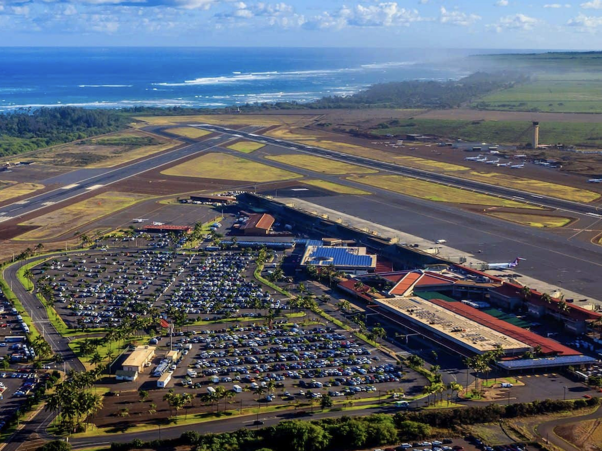 The image shows an aerial view of an airport near the coastline, featuring runways, terminals, a parking area, and open fields.