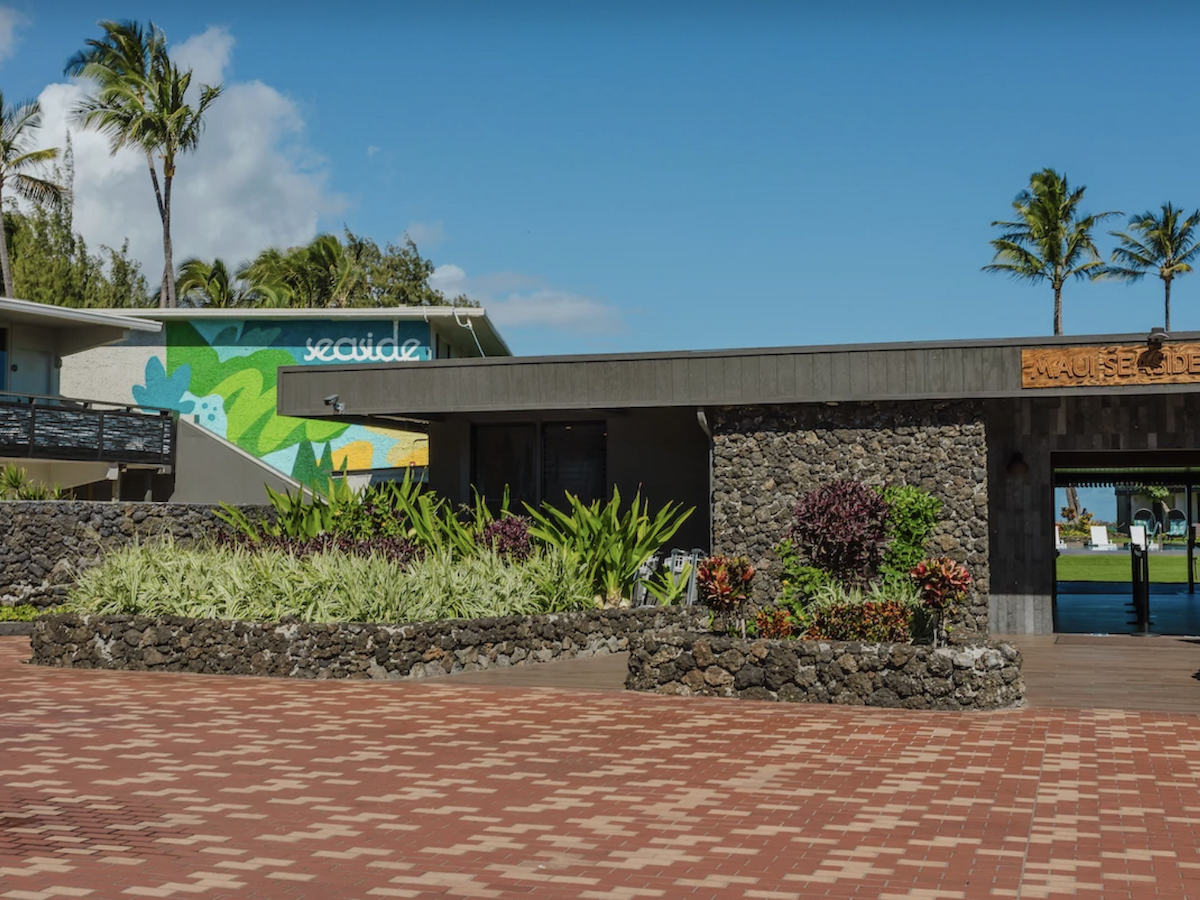 The image shows a modern building with stone accents, tropical landscaping, and a mural. Palm trees and clear skies are in the background.
