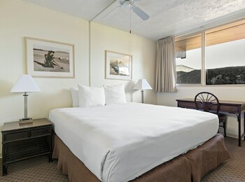 The image shows a hotel room with a bed, two lamps, two framed pictures, a desk, chair, and a window with blinds.
