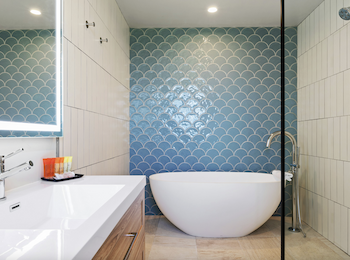 A modern bathroom with a freestanding tub, blue scallop-tiled wall, white vanity with a mirror, and a glass-walled shower.