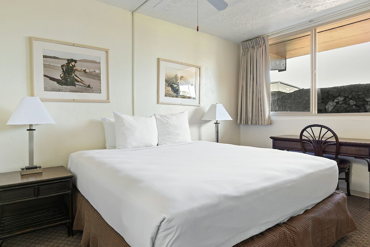 A hotel room with a neatly made bed, two lamps, framed wall art, a window, and a desk with a chair.