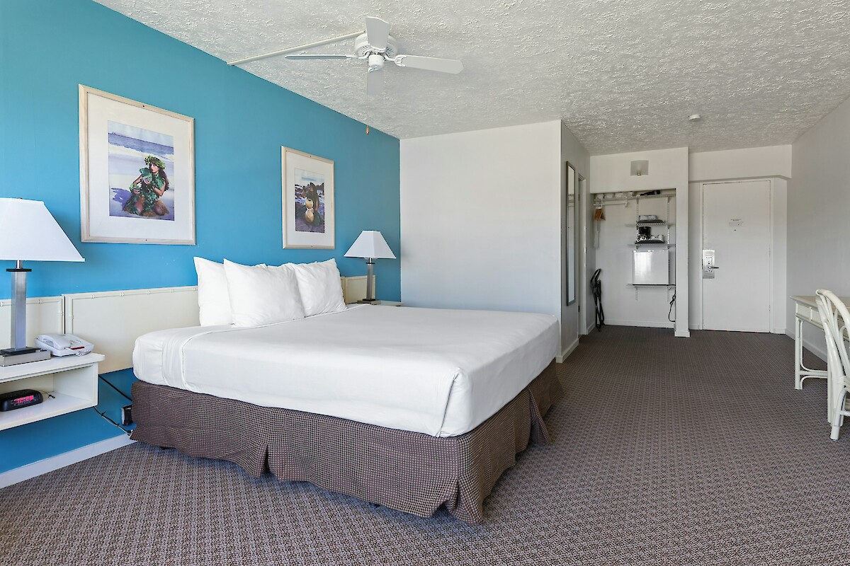 A hotel room with a bed, blue accent wall, two lamps, a small desk, and a closet area in view. The decor is simple and clean.