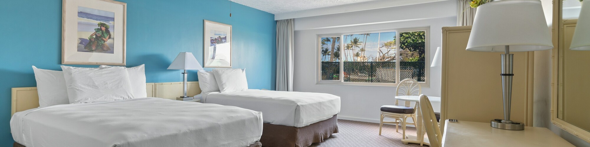 A hotel room with two beds, a blue accent wall, artwork, a desk, lamps, and a window showing a view of palm trees.